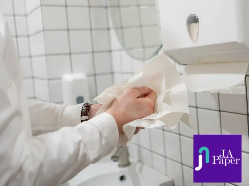 Pessoa secando as mãos com papel interfolha em um banheiro comercial, com dispenser de papel e azulejos brancos ao fundo. Ideal para empresas que priorizam higiene e praticidade.
